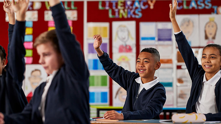 Three primary school students sitting in a classroom with hands raised in participation.
