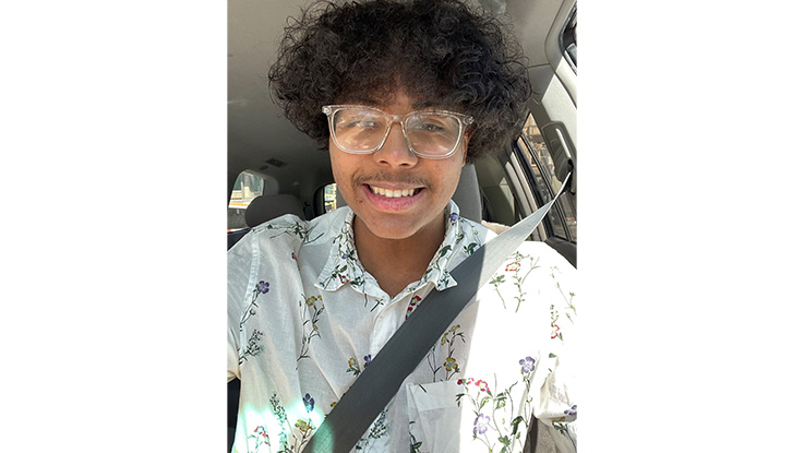 a boy sitting in a car with his seat belt on wearing glasses and wearing a light blue short sleeve shirt smiling