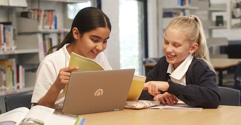 two school girls in a library with a laptop working on a project together